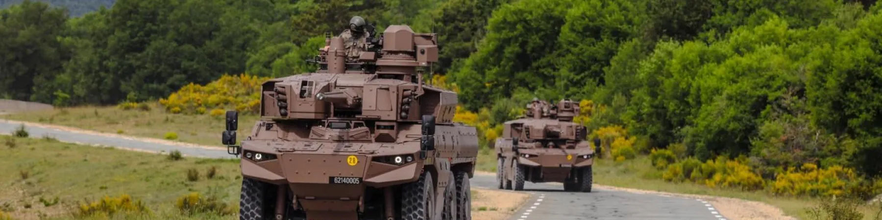 Two brown armored vehicles on the road. Man observing from the front turret.