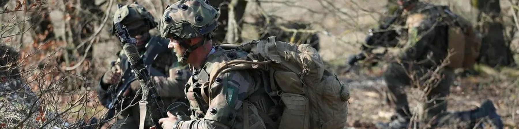 Soldats en treillis et casques, à genoux ou accroupis, en position tactique dans un environnement boisé et sec, avec fusils.